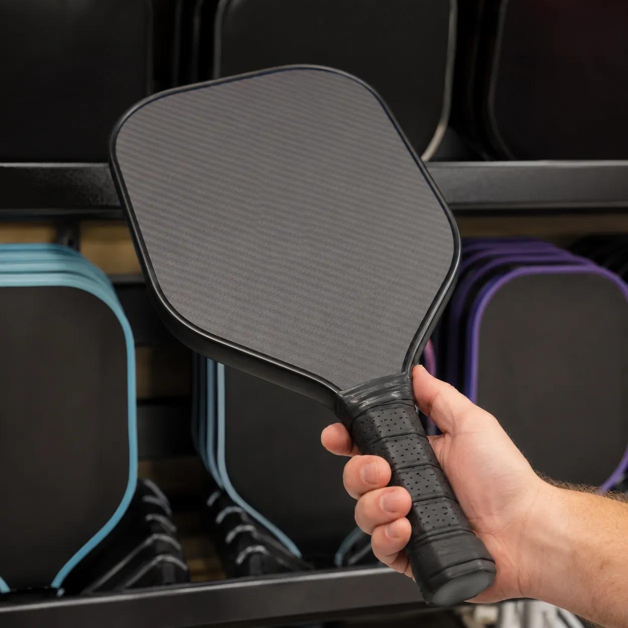 A bare pickleball paddle face and handle held in front of a retail rack, the product surface Article 9(6) reaches for before the identification block may move to the packaging