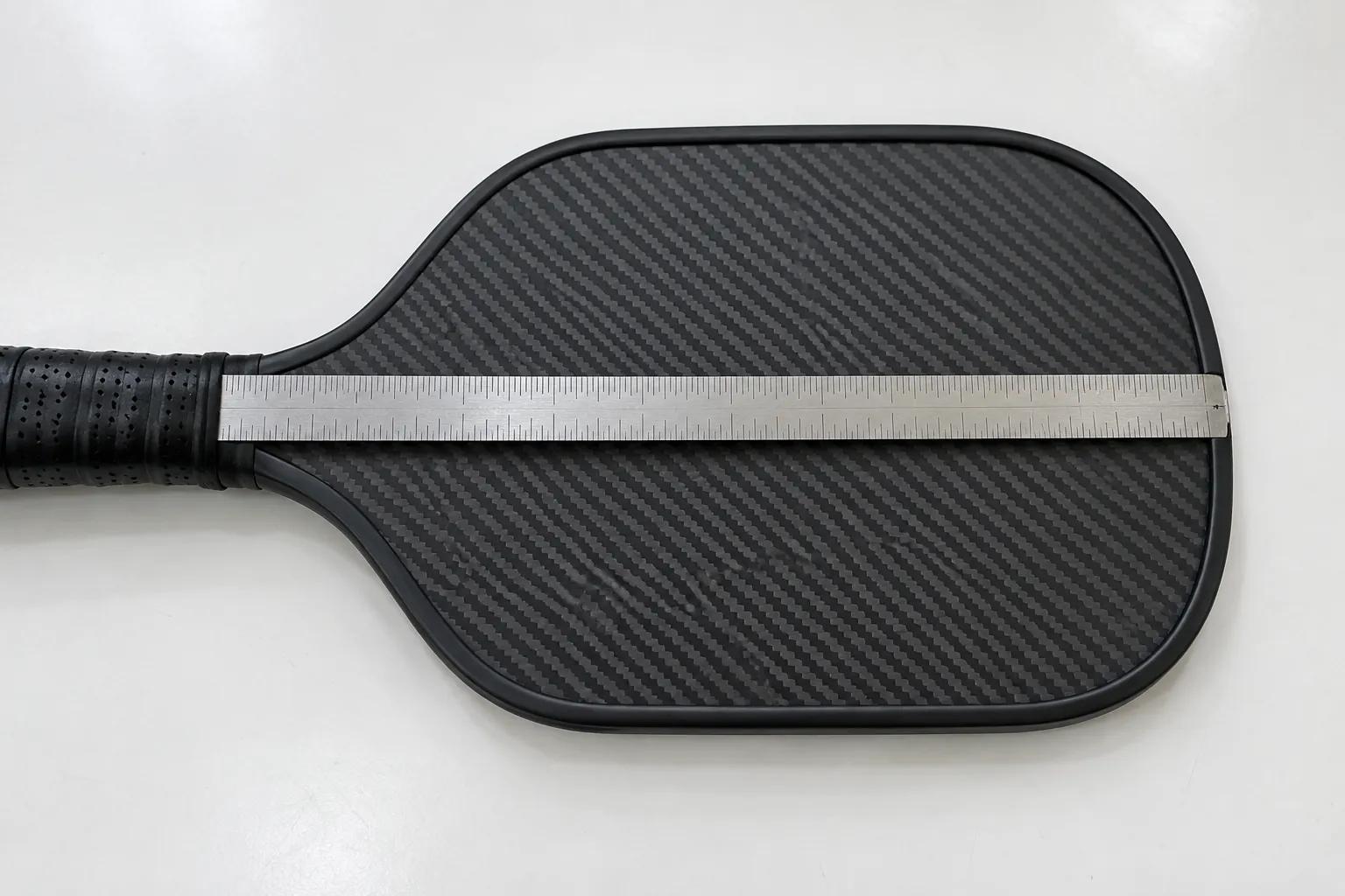 A steel rule laid across a blank carbon fiber pickleball paddle fitted with a black edge guard, the end of the rule at the outer edge of the guard