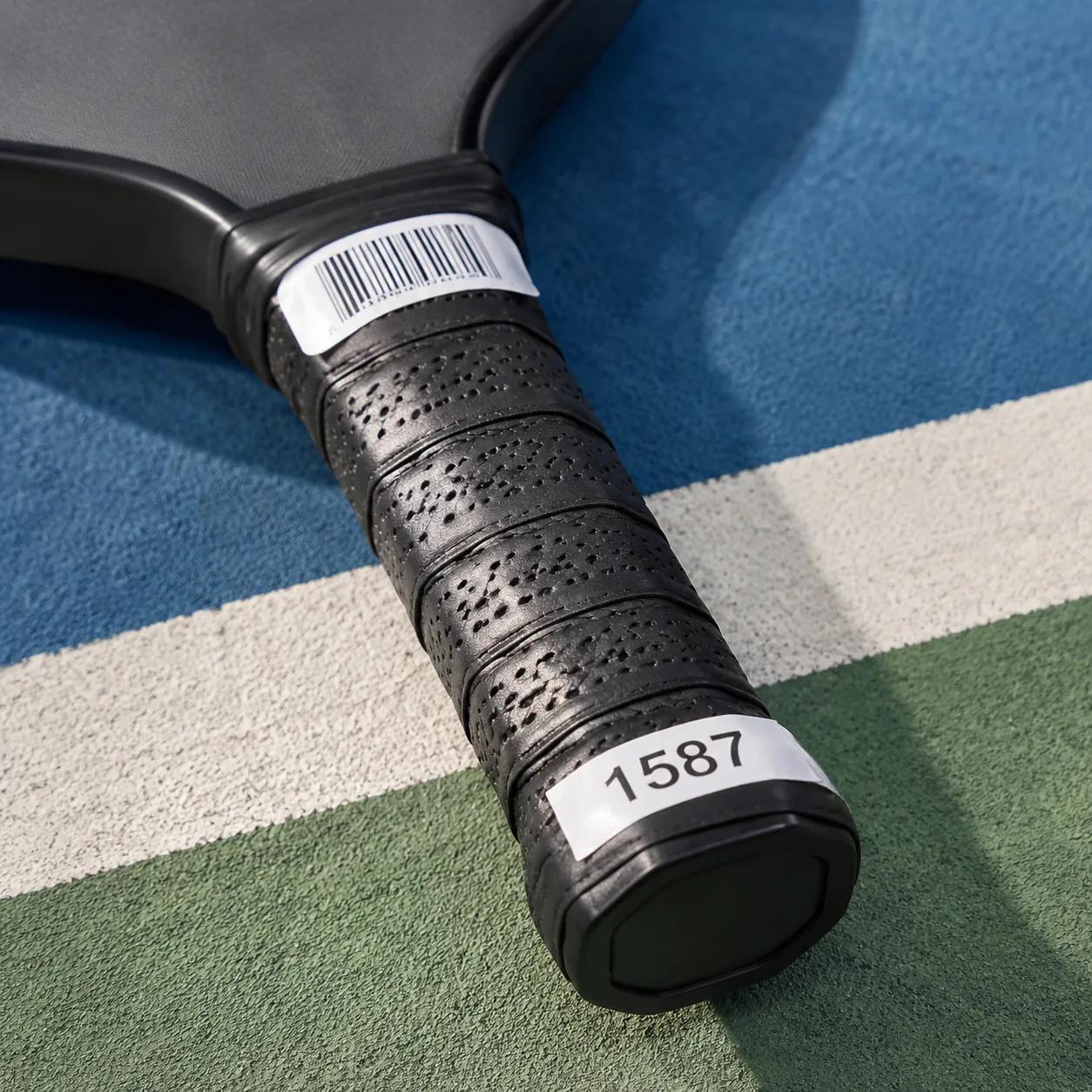 Hyper-realistic product photography, close-up of a pickleball rental paddle handle with a durable barcode sticker and numbered fleet tag, textured grip wrap, natural daylight, clean product shot, no text, no brand logo