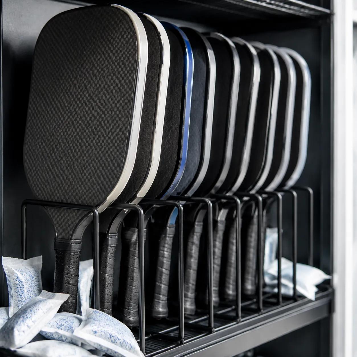 Hyper-realistic product photography, a row of pickleball paddles stored vertically in a cool, dry storage rack with silica gel packets nearby, soft ambient light suggesting a controlled environment, clean composition with shallow depth of field, focus on paddles and moisture-absorbing accessories, no text, no brand logo