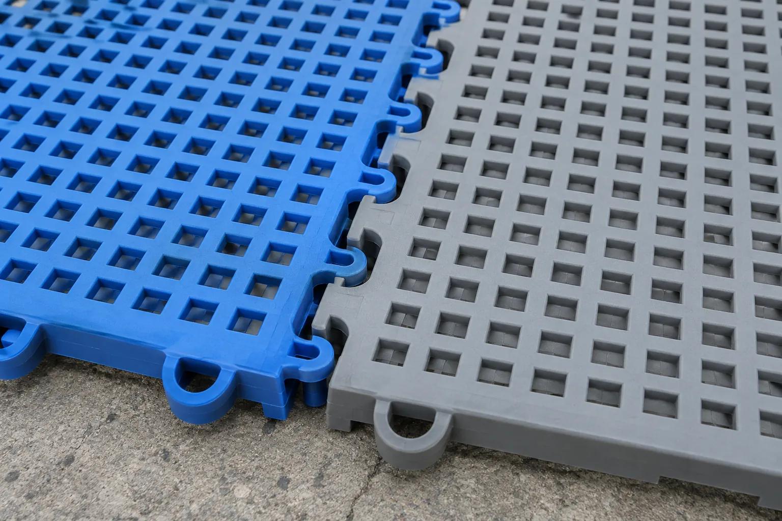 Close-up of blue and grey modular interlocking court tiles laid over a concrete slab, showing the moulded open grid of the tile face and the loop interlock joining two tiles