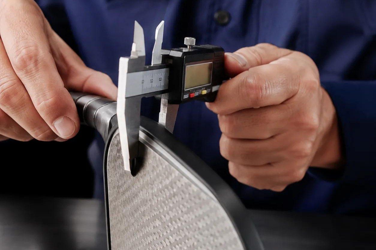 A digital caliper closed across the edge guard of a carbon-fibre pickleball paddle held in two hands