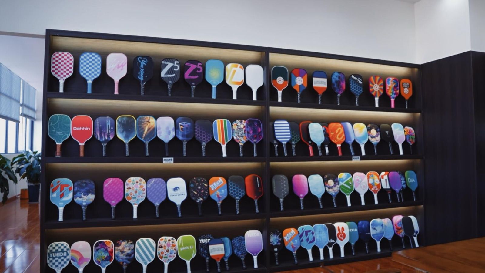 Factory showroom wall of finished pickleball paddles in full-colour printed face artwork, showing the branding freedom a painted face allows