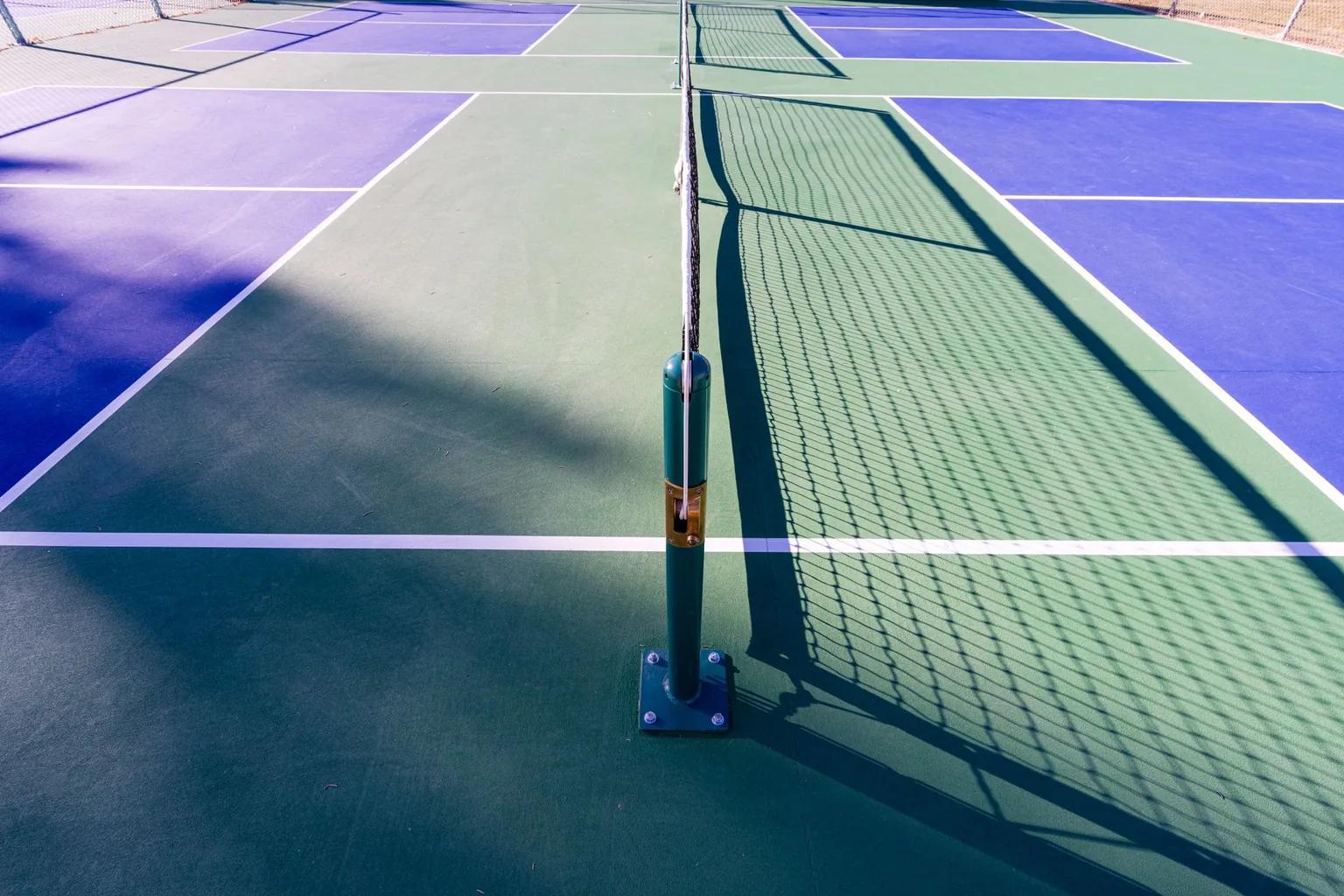 Net post on a pickleball court bolted to the acrylic colour coat through a square steel base plate, the net casting a shadow across the green and blue surface