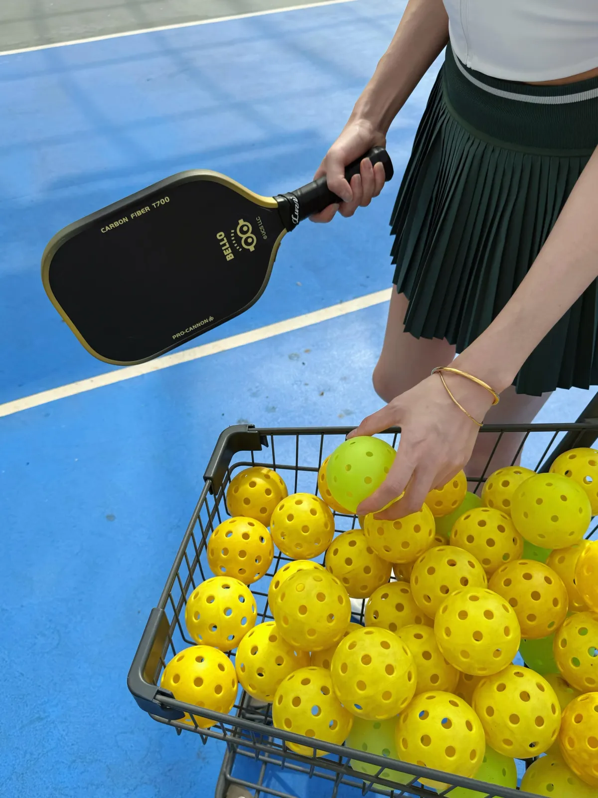 Hand selecting a yellow pickleball from a wire basket beside a paddle