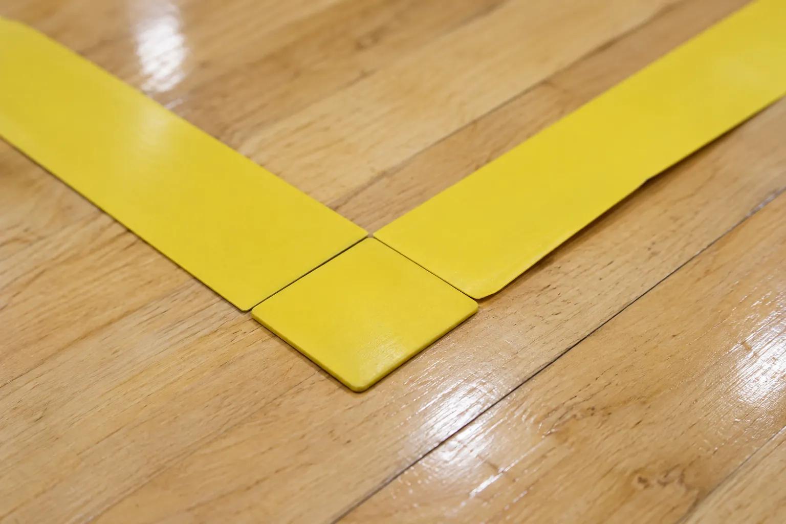 Close-up of a square yellow corner piece butted against two flat yellow line strips on a varnished maple floor, with a thin seam at each joint and one strip edge lifting slightly