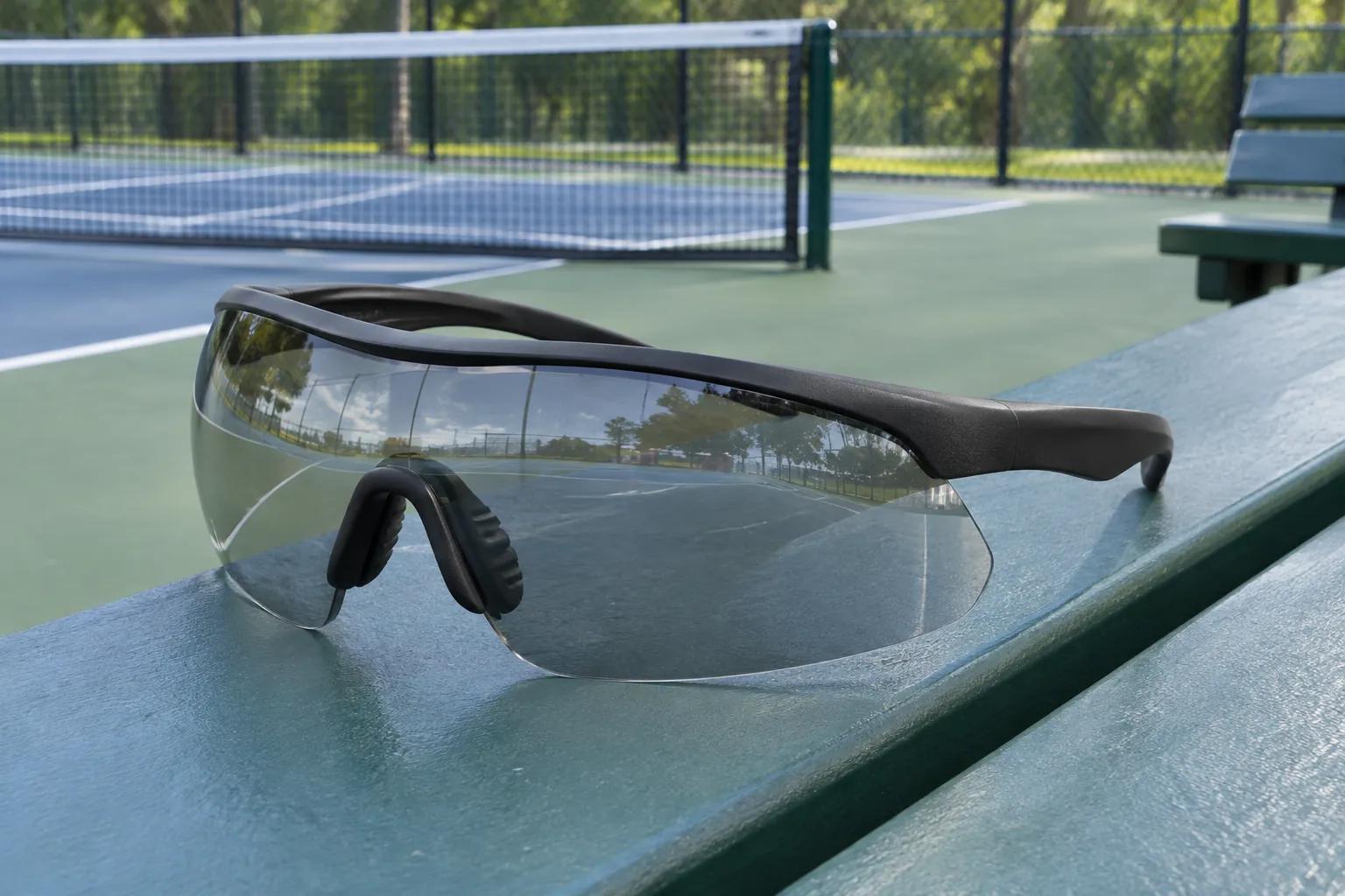 Clear-lens wraparound sports eyewear with a one-piece curved lens and a plain black frame resting on a courtside bench beside an outdoor pickleball court