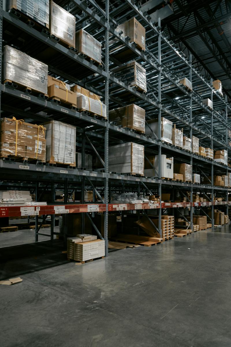 Hyper-realistic commercial photography, hundreds of identical pickleball paddles stacked in neat rows on wooden pallets inside a large warehouse, warm ambient lighting from overhead industrial fixtures, wide angle composition showing the massive scale of bulk inventory, subtle dust particles in the air, no text, no brand logo