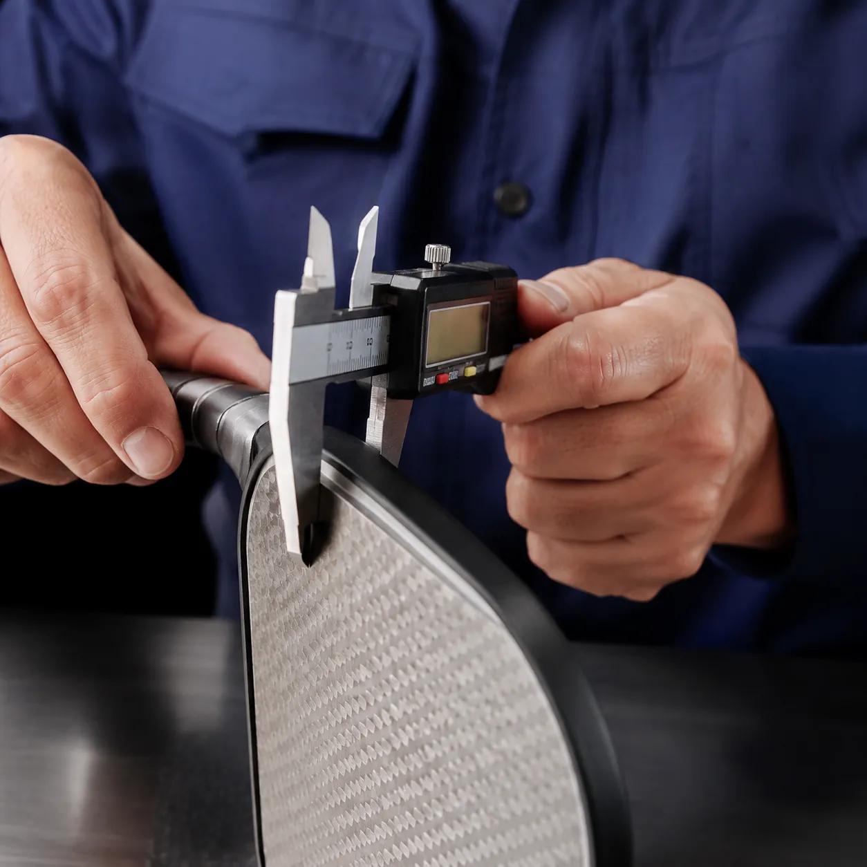 Hyper-realistic product photography, a quality control inspector using a digital caliper to measure a pickleball paddle thickness, close-up of hands and paddle, precise workshop lighting, professional accuracy, no text, no brand logo