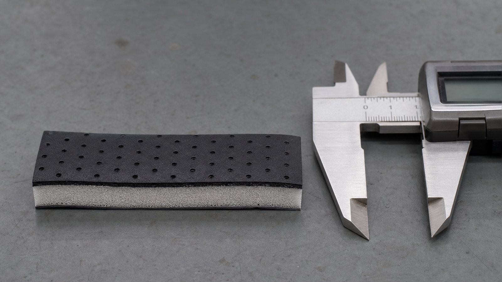 Cut section of a pickleball replacement grip showing a perforated black polyurethane top skin bonded to a pale EVA foam carrier, with a digital caliper alongside