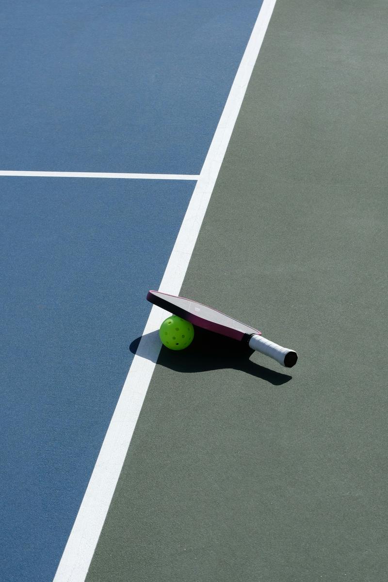 Hyper-realistic product photography, single prototype pickleball paddle resting on a workbench next to technical specification documents and material swatches including carbon fiber weave samples, workshop environment, focused spotlight lighting, rapid prototyping aesthetic, no text, no brand logo