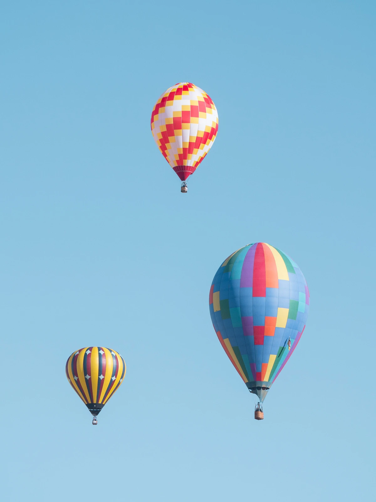 Three hot-air balloons — a red-and-white checker, a multicolored patchwork, and a yellow-striped one — floating against a clean blue sky