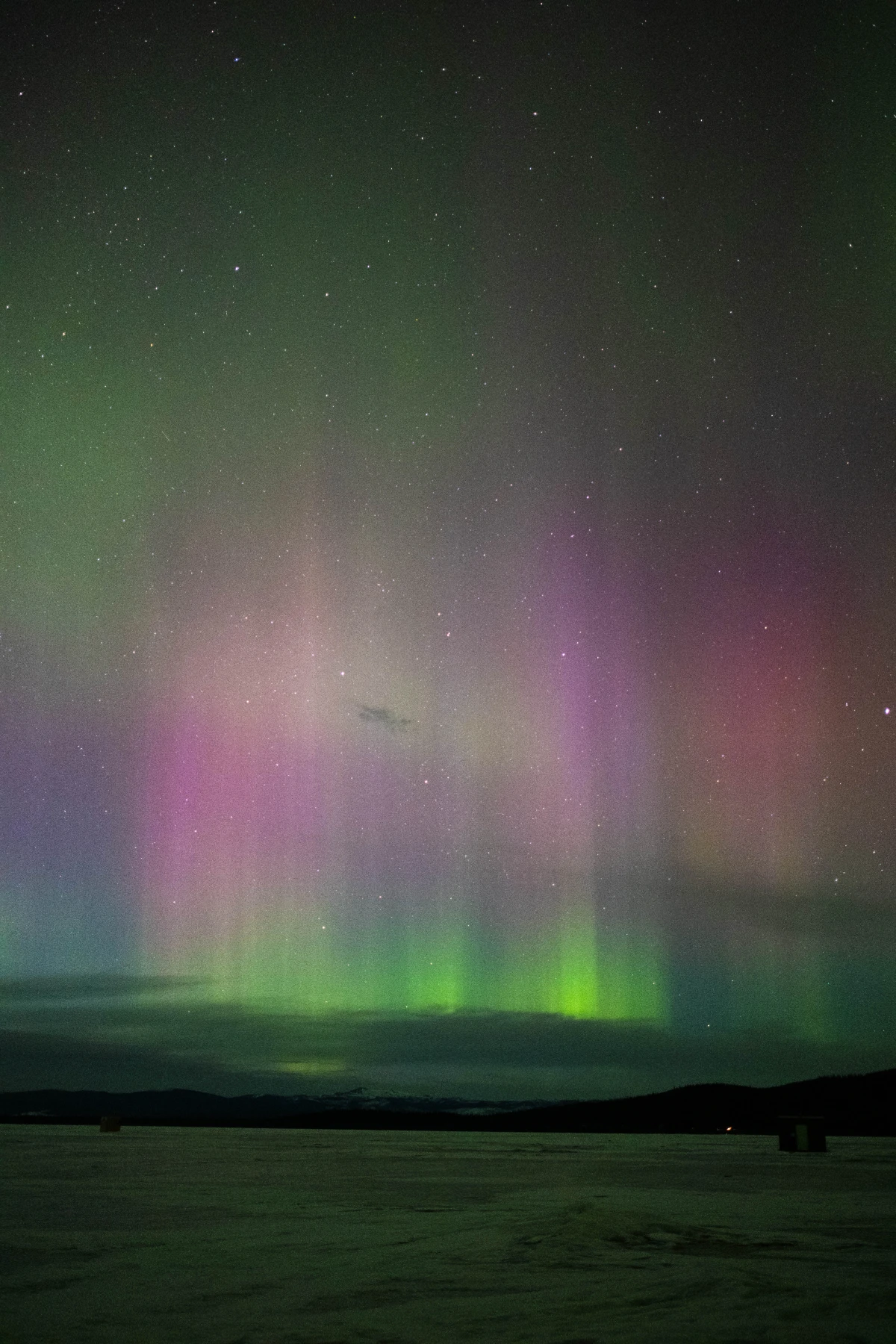 Green and pink aurora pillars reflected over a frozen lake with small ice-fishing huts on the horizon