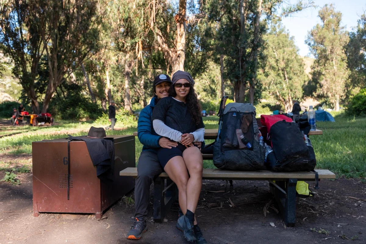 Two backpackers sitting close together on a wooden picnic table at the campground, packs and a metal food-storage box beside them, eucalyptus trees behind
