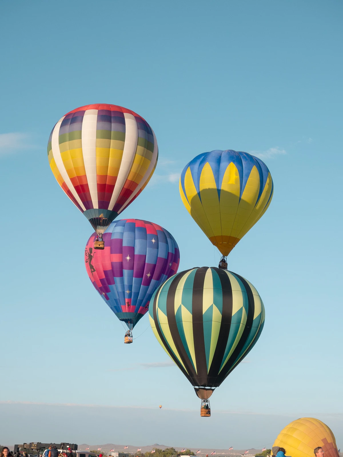 Four hot-air balloons hanging low over the field above flags and a gathered crowd at sunrise