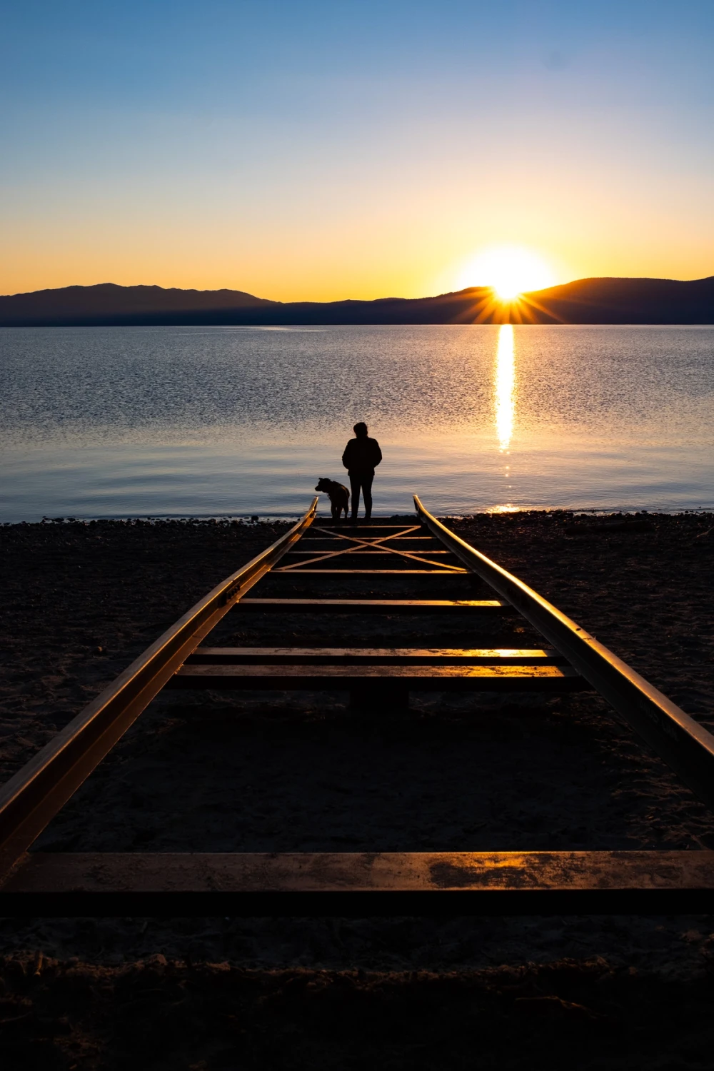 Silhouette of a person and a dog standing at the end of old wooden boat tracks leading into Lake Tahoe at sunset