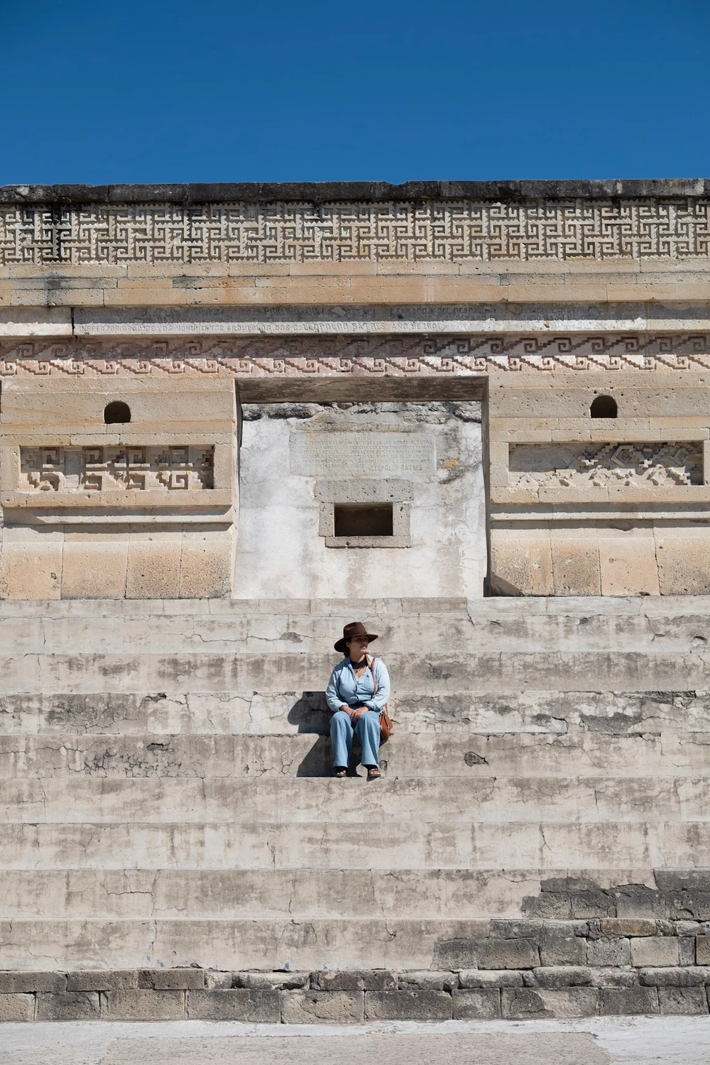 A person in a hat sitting on the broad stone steps of a Mitla building below a frieze of geometric fretwork against a deep blue sky
