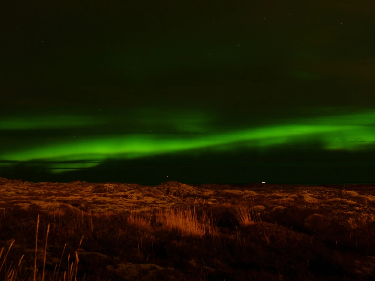 Green northern lights arcing across a dark sky above a faintly lit Icelandic lava field