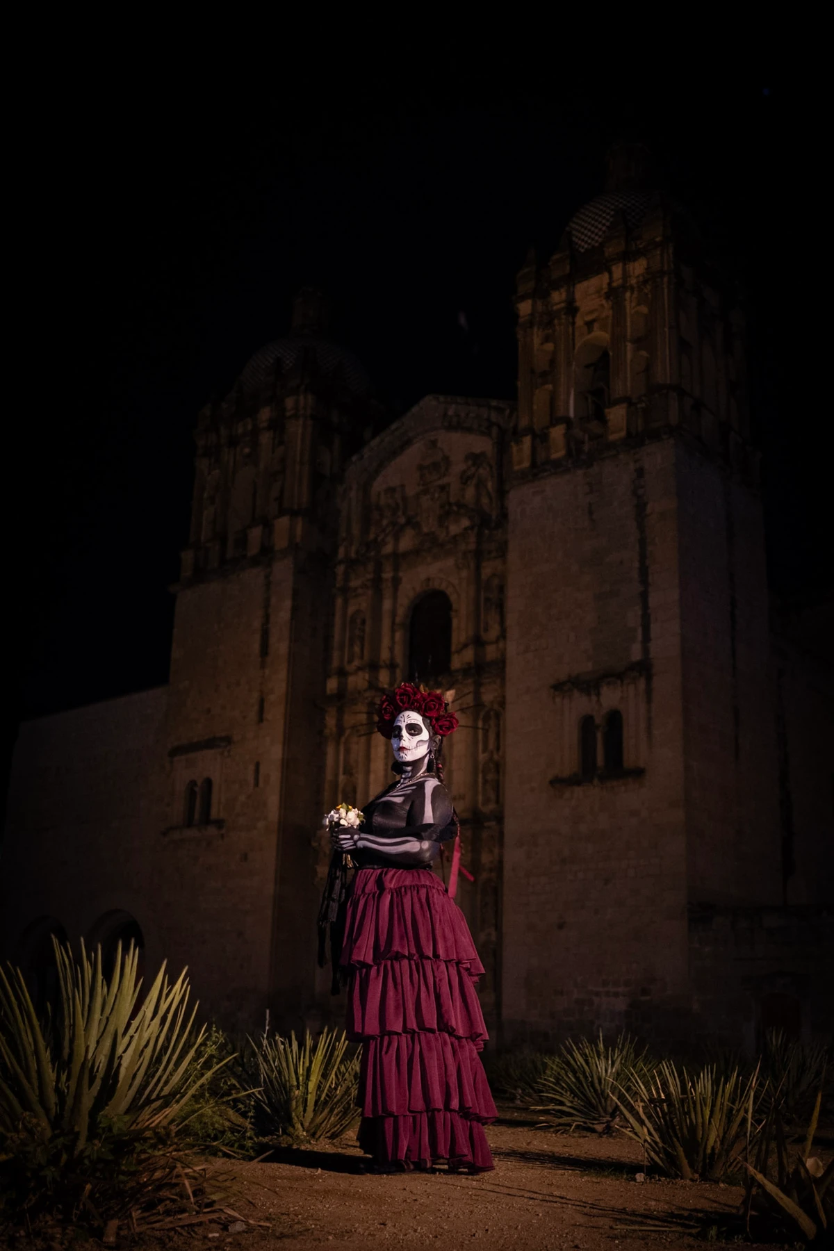 A Catrina in a tiered deep-red dress and flower crown stands among agave plants before the floodlit baroque facade of the Templo de Santo Domingo at night