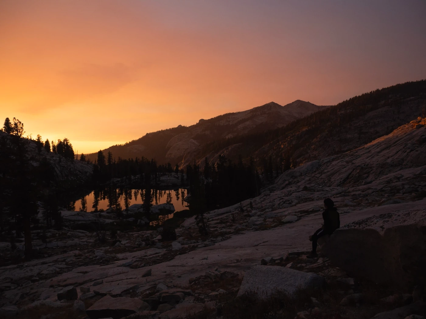A hiker sitting on a granite slab at dusk above a small alpine lake, distant peaks glowing peach under a smoky sky