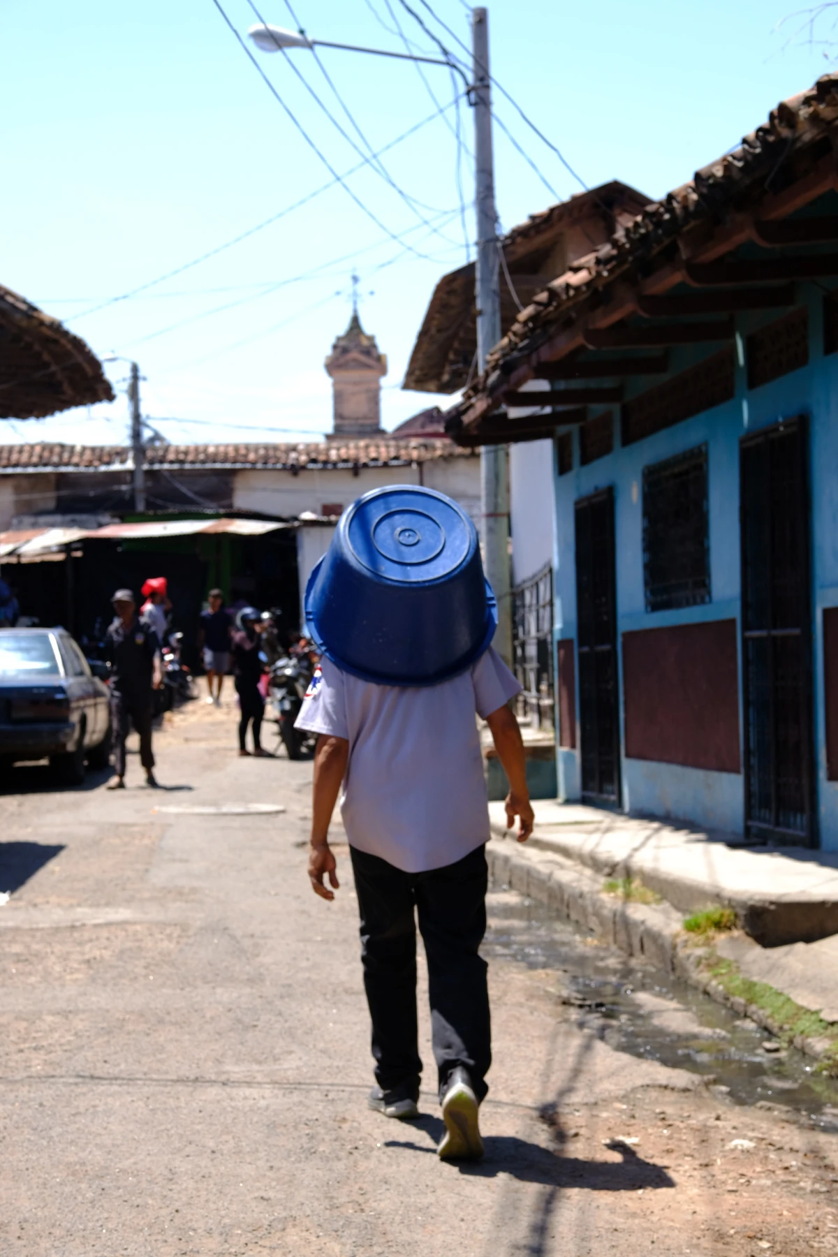 A person carrying a large blue water jug on their shoulder down a street of terracotta-tiled rooftops, a church tower in the distance