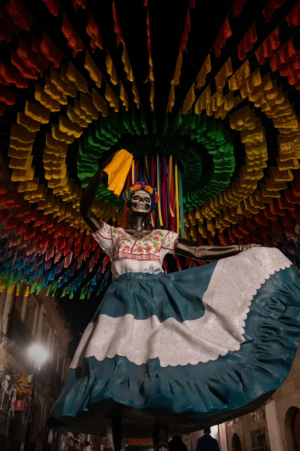 A towering Catrina puppet in a traditional teal-and-white Oaxacan dress, photographed from below beneath an arched tunnel of colorful papel picado