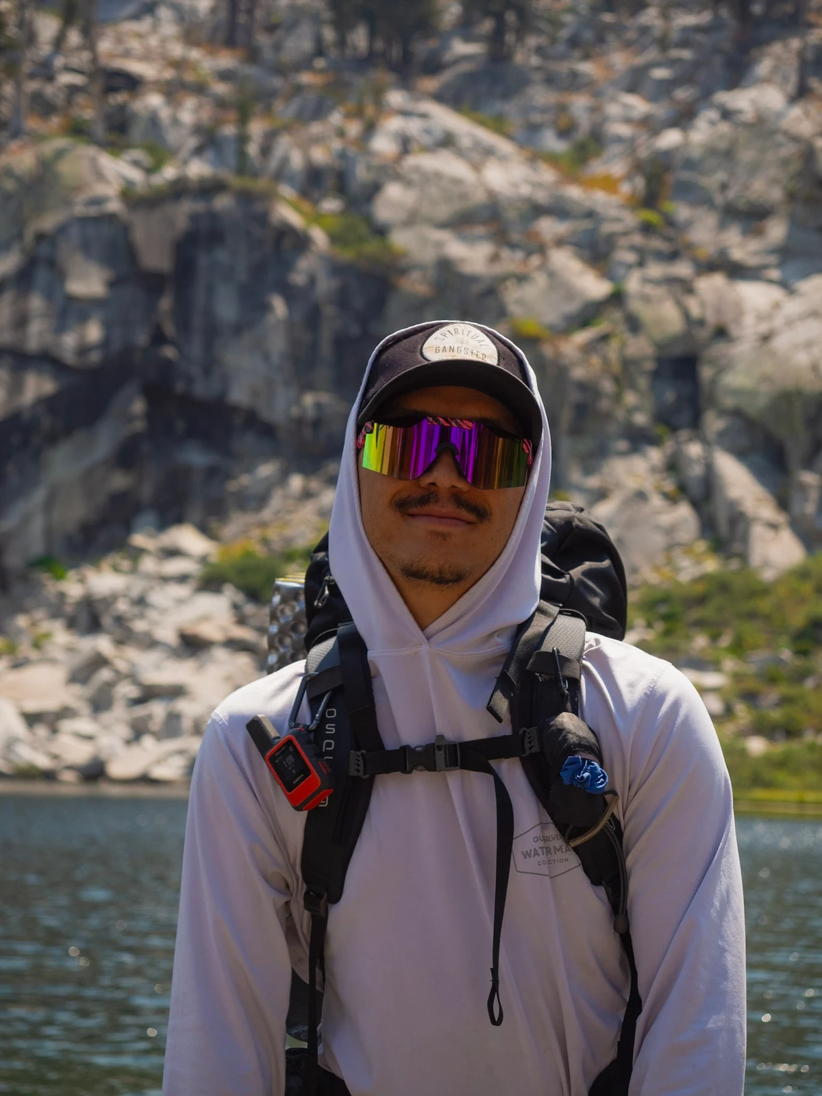 A hiker in a white sun hoodie, brimmed cap, and mirrored sunglasses standing in front of a granite-walled lake with a loaded backpack