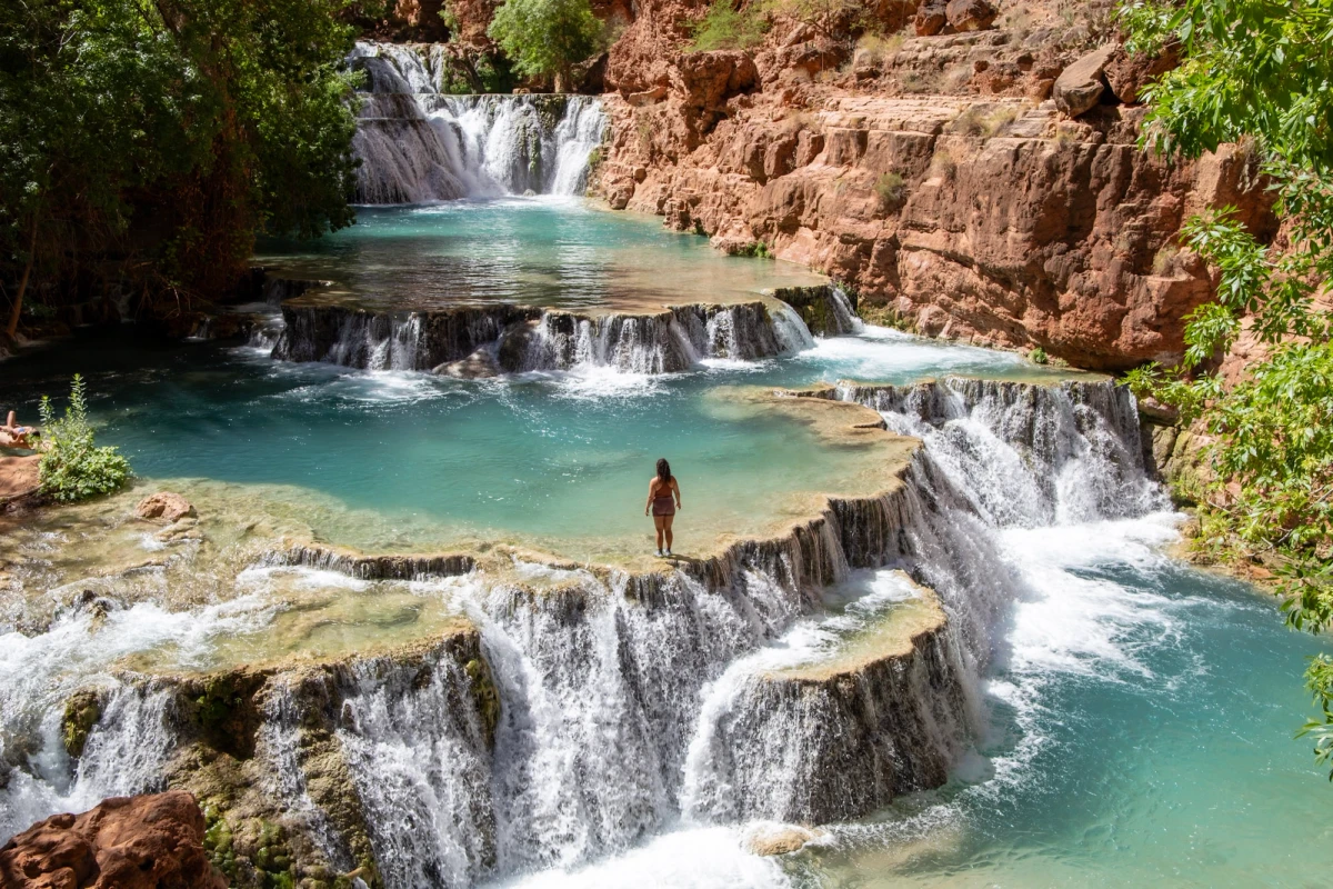 Tiered turquoise pools and low travertine waterfalls at Beaver Falls, a person standing on a ledge between them among red walls and greenery