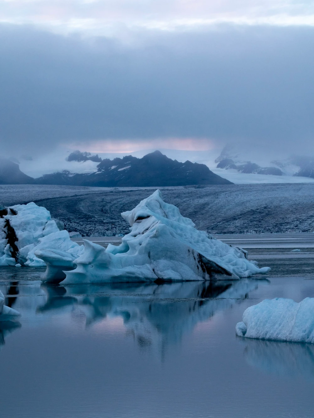 Icebergs reflected in the still Jökulsárlón lagoon at dusk with the glacier tongue and clouds behind