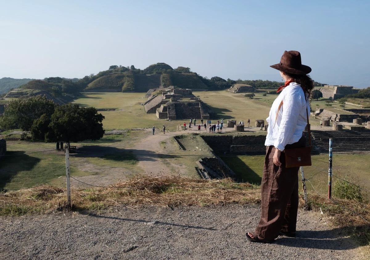 A figure in a wide-brimmed hat and shoulder bag stands on a rise overlooking the broad grass plaza and stone platforms of Monte Albán