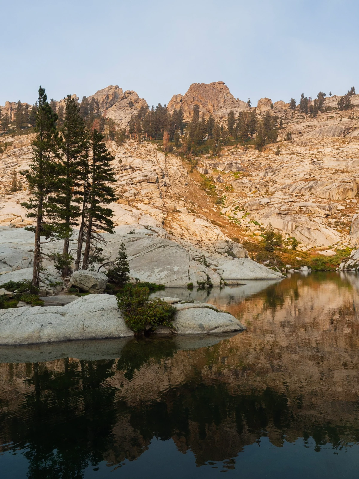 Two tall pines on a granite slab beside a still lake in soft morning light, with jagged granite peaks reflected in the water