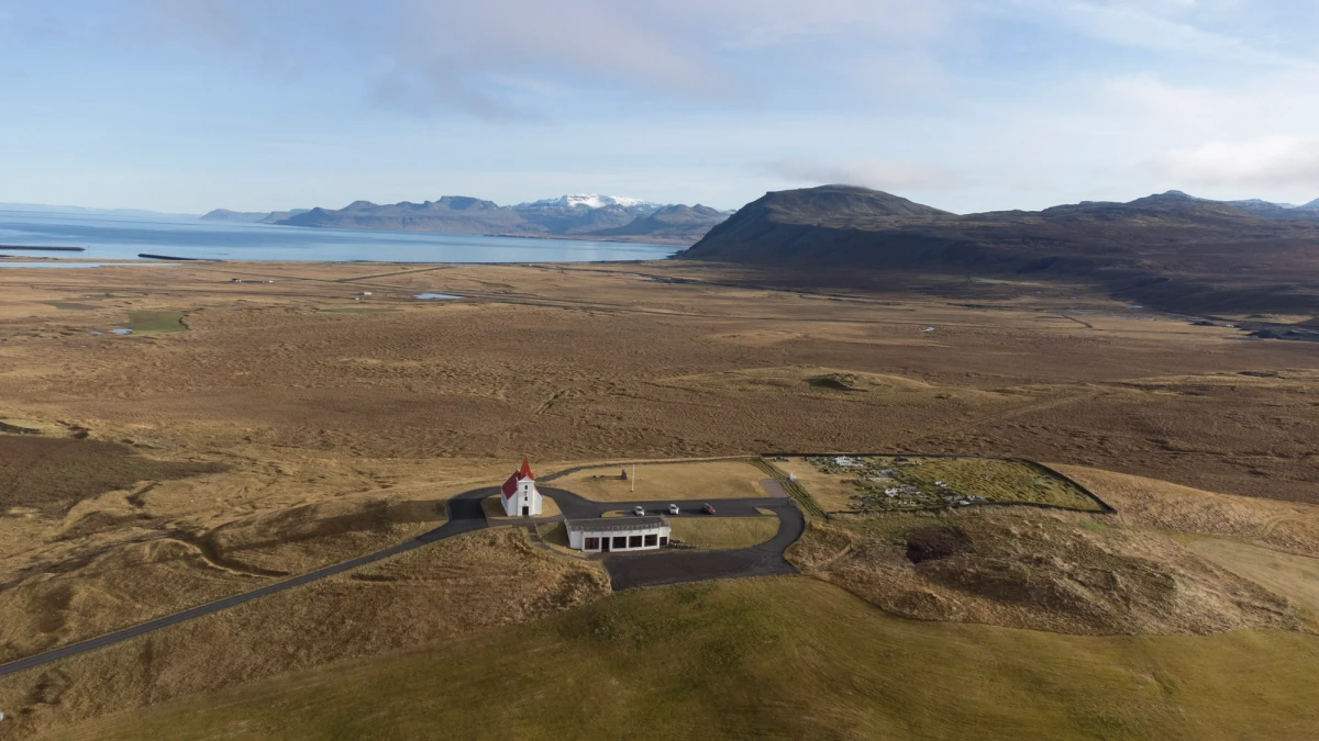 Aerial view of a small white-and-red Icelandic church alone on a golden plain above a fjord, mountains beyond