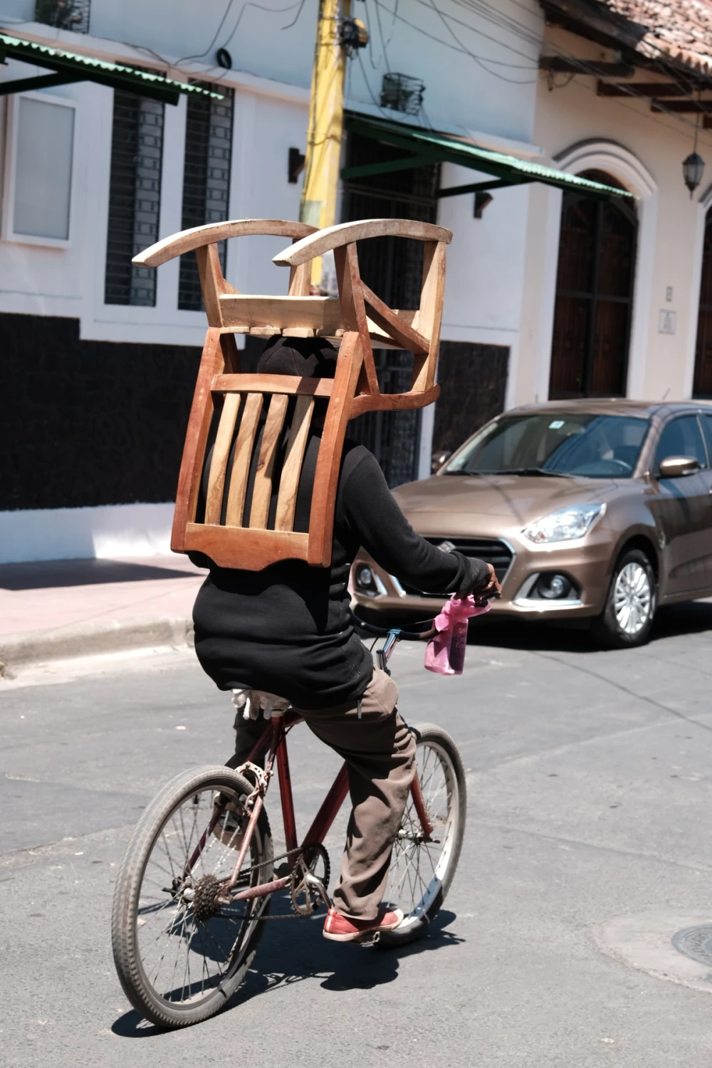 A person cycling down a sunlit street with a wooden chair balanced on their back, colonial buildings behind