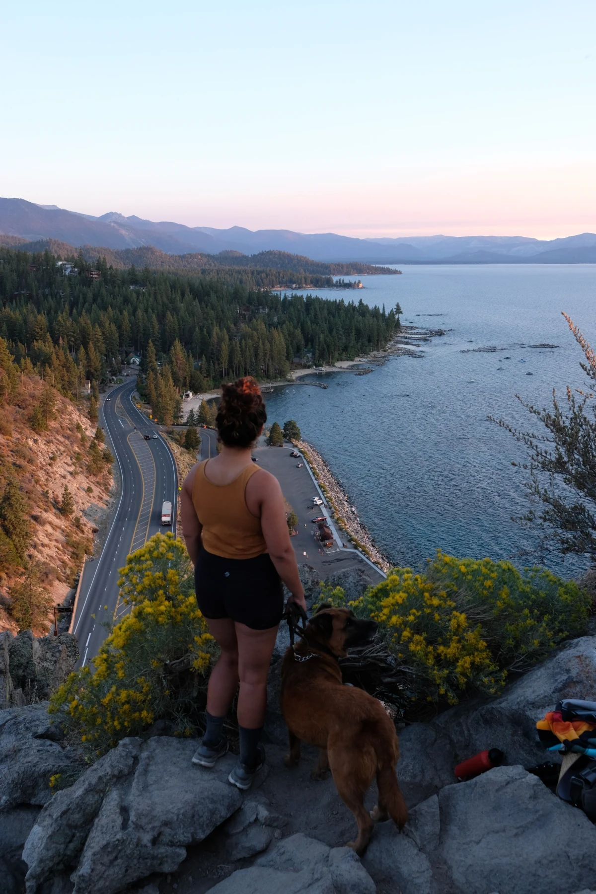 A person and a dog resting on rocks above a Lake Tahoe cove at golden hour, a winding shoreline road below