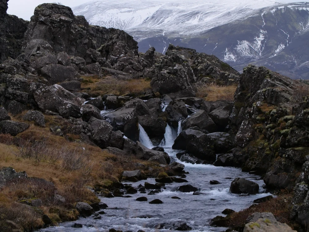 A small waterfall dropping through dark lava rock with a snow-covered mountain behind