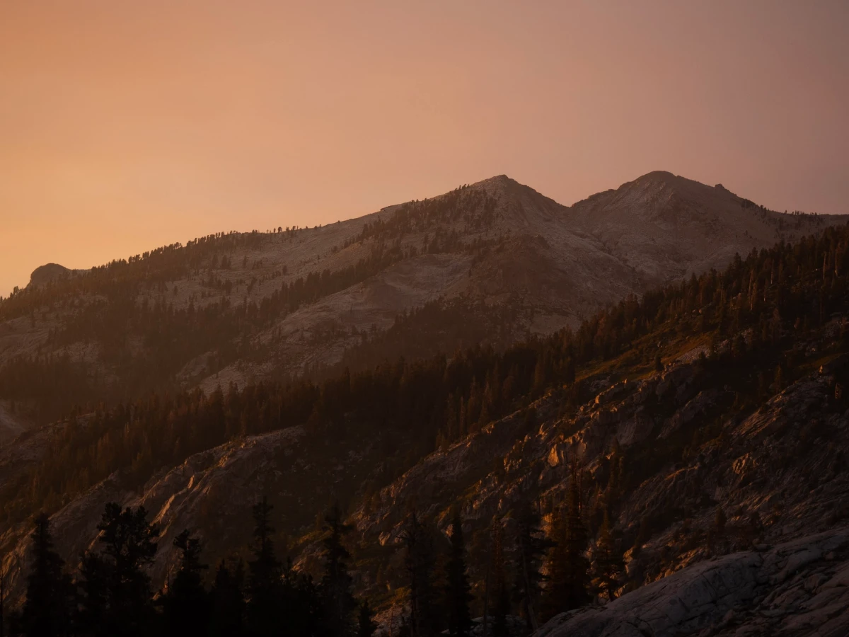 Layered granite ridgelines fading into peachy wildfire haze at sunset, dark forested slopes below