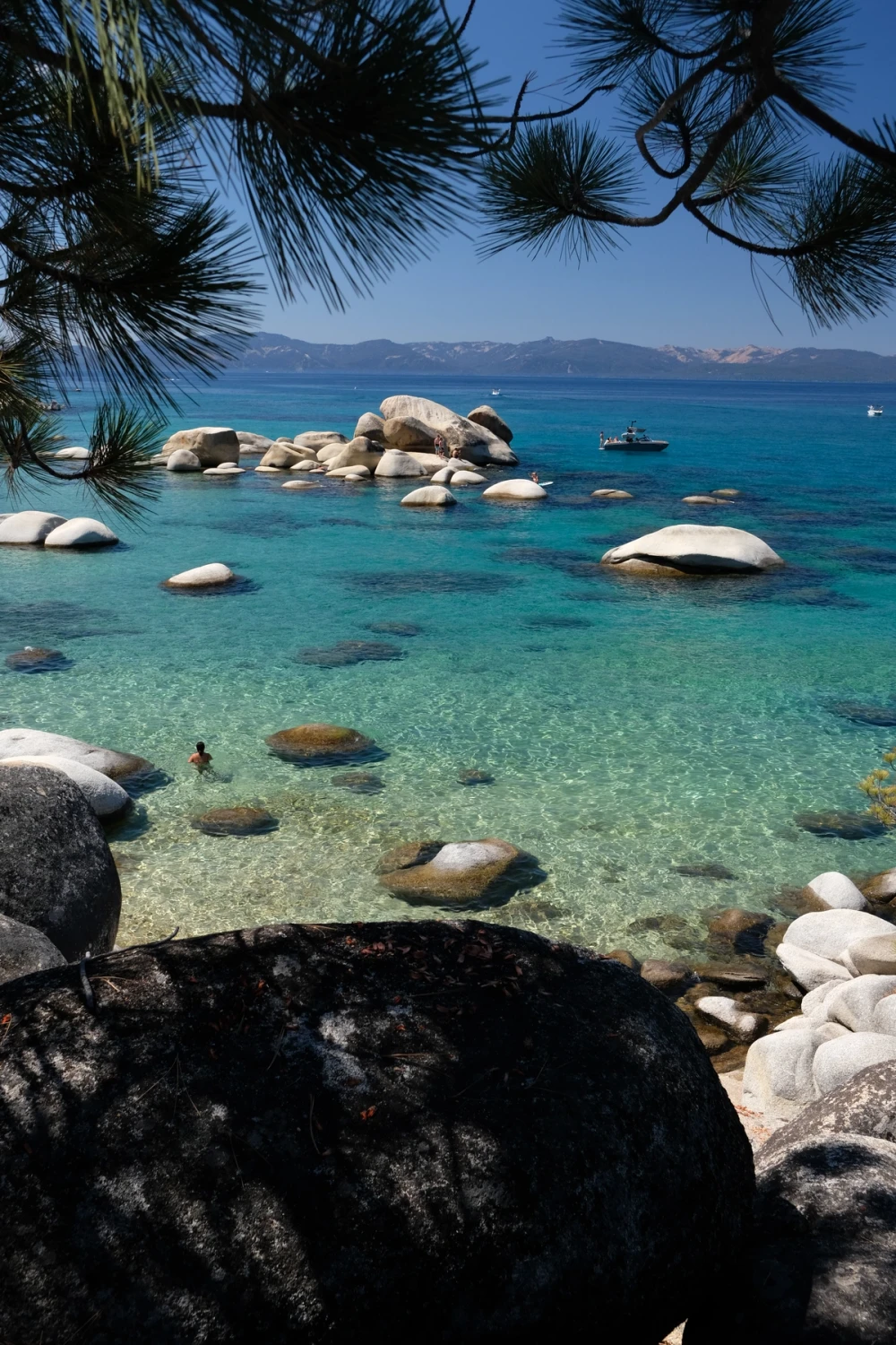 Crystal-clear turquoise water over scattered granite boulders along the Lake Tahoe shore, framed by a boulder and pines