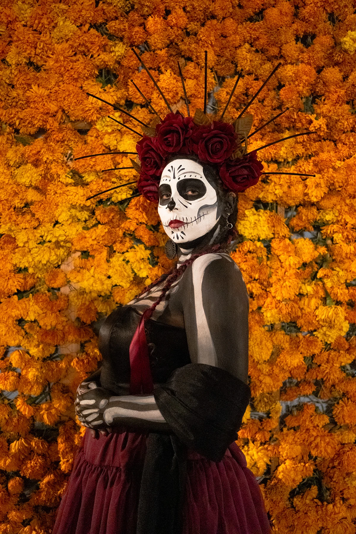 A woman in detailed Catrina makeup and a crown of red roses and gold spikes, posed against a dense wall of orange marigolds