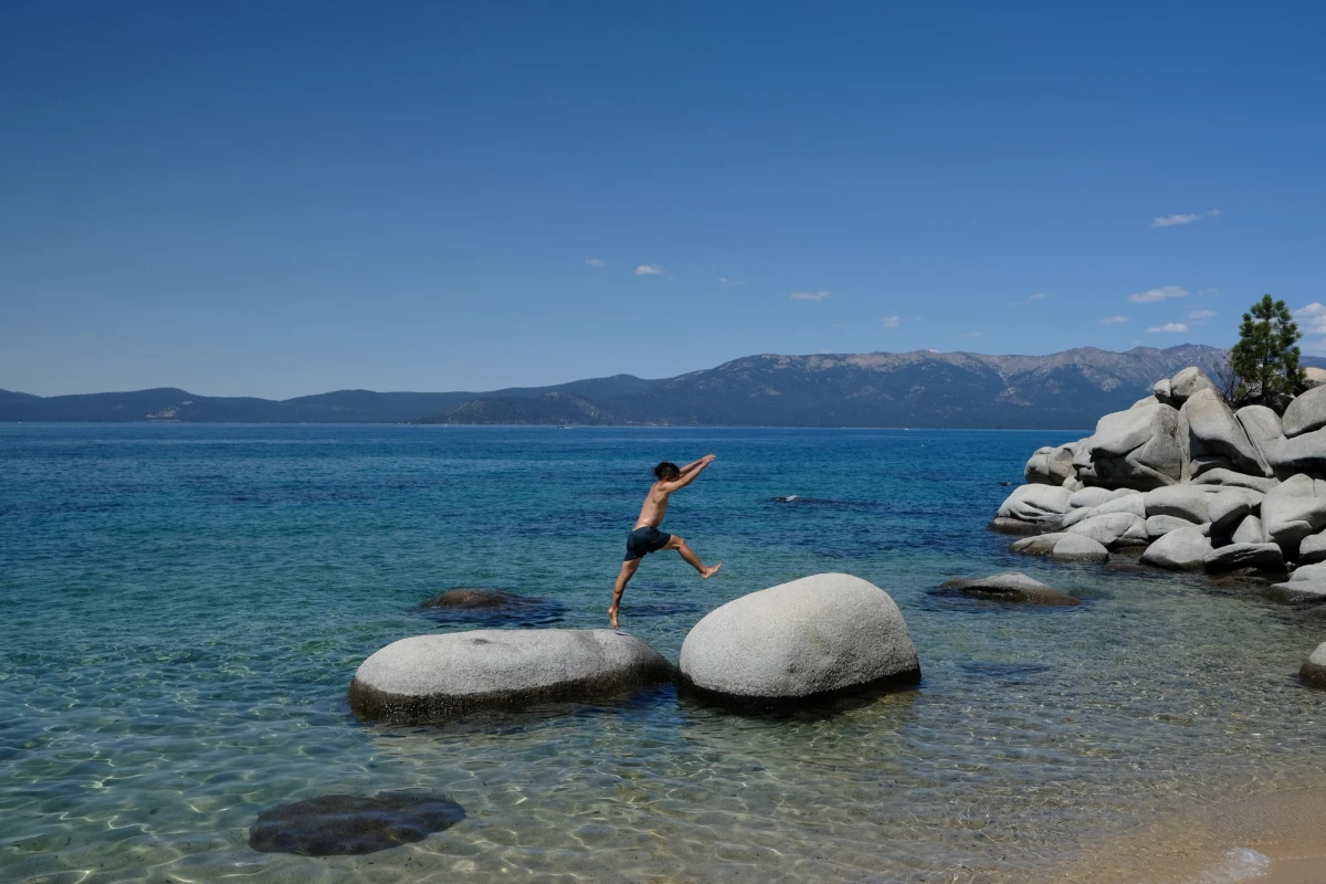 A person leaping between two large granite boulders in the crystal-clear shallows of Lake Tahoe, mountains and blue sky behind