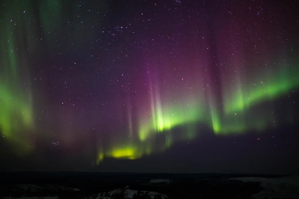 A broad green and magenta aurora band stretching horizon to horizon over a dark Alaskan valley