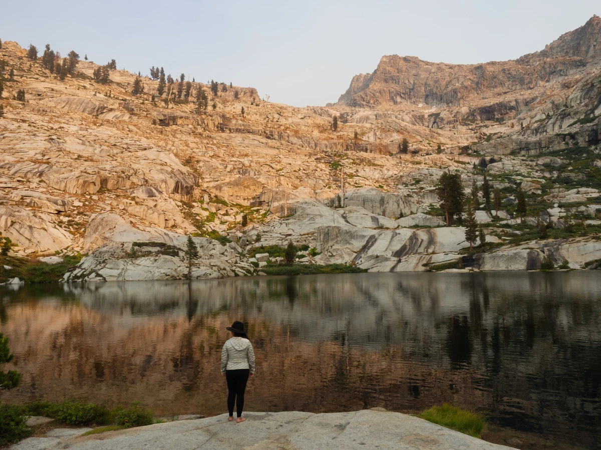 A person standing on a granite slab at the edge of a calm lake, looking up at a sunlit granite mountainside mirrored in the water