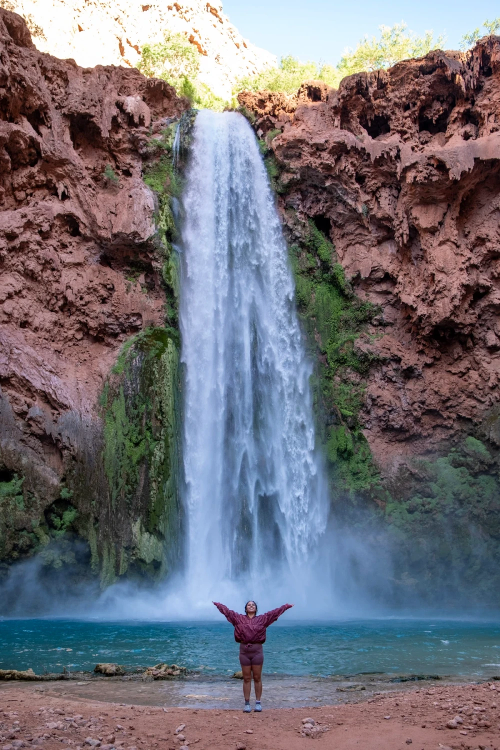 A person standing with arms outstretched at the base of Mooney Falls as the waterfall drops into a blue pool