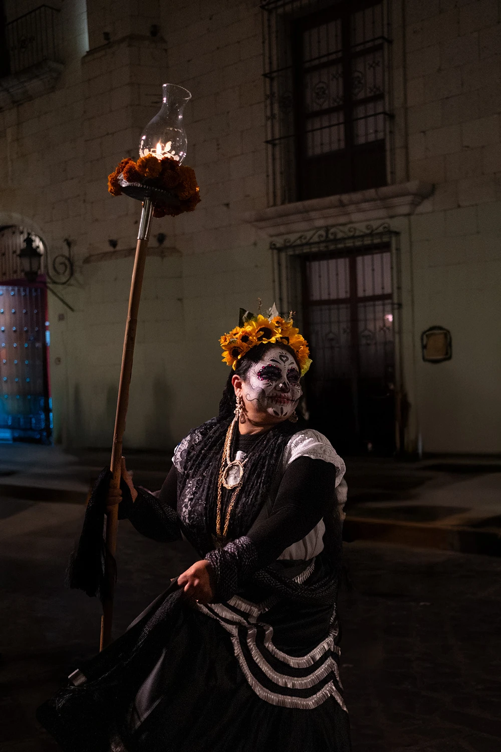 A woman in Catrina makeup and a sunflower crown, in a black-and-white ruffled dress, holding a tall staff topped with a glowing lantern and marigolds