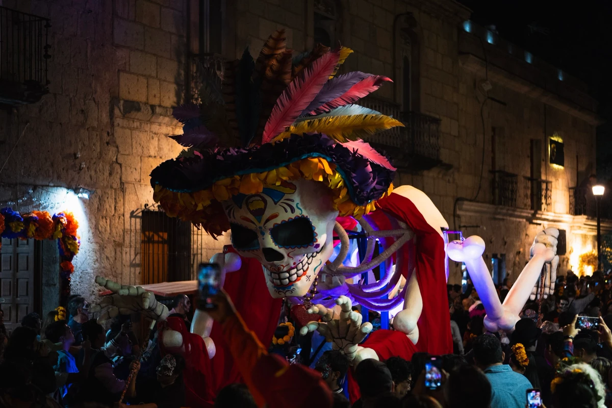 A giant illuminated Catrina mojiganga puppet with a feathered hat towers over a crowd of phones on a Oaxaca street at night during Día de los Muertos