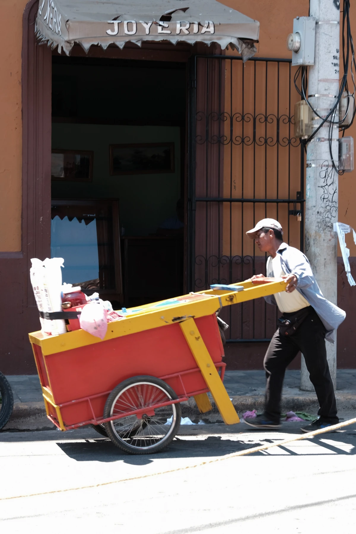 A vendor pushing a red-and-yellow wooden cart past a jewelry shop with iron grillwork and an ochre colonial wall in bright sun