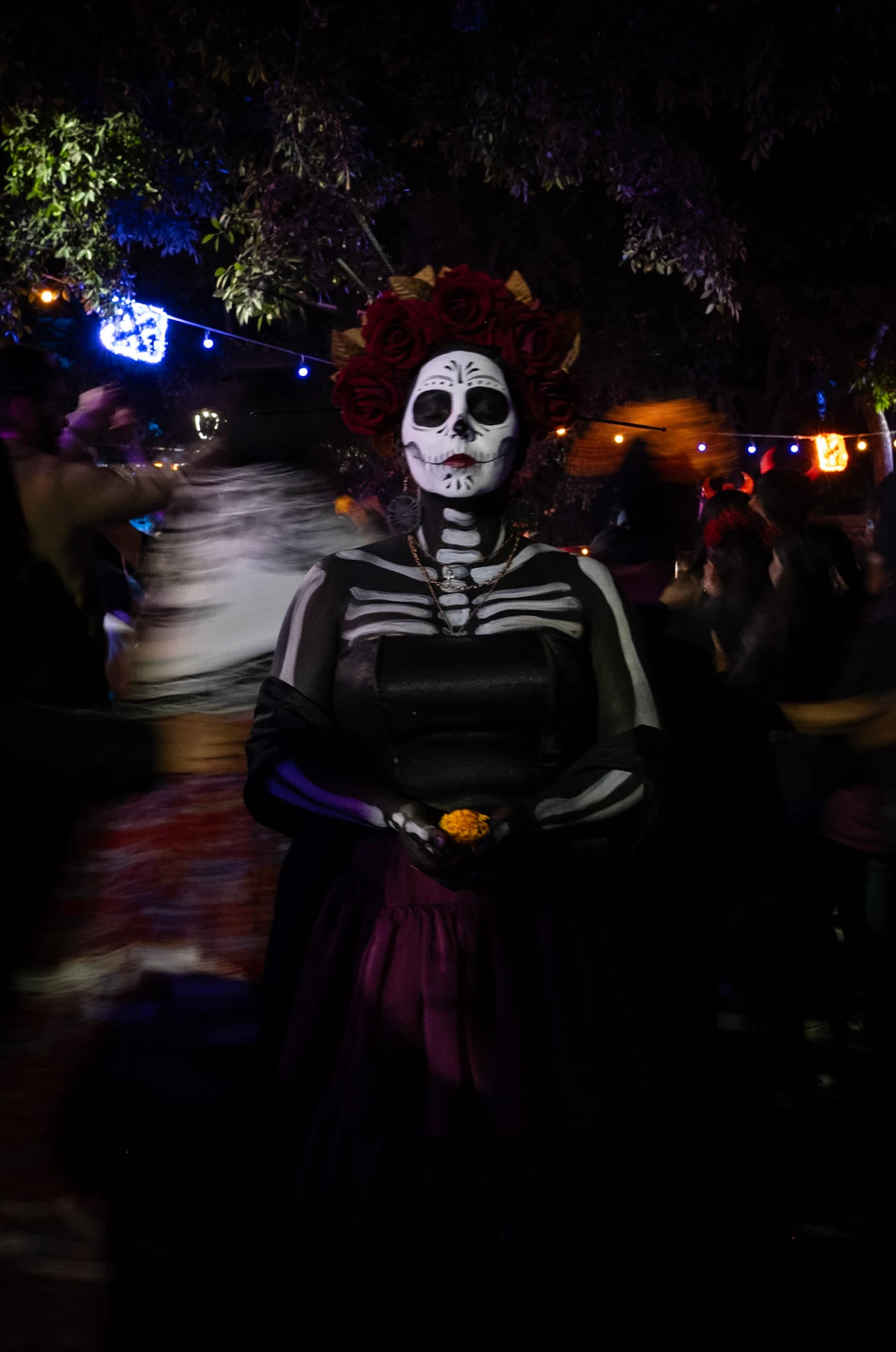A woman in white Catrina face paint and a red rose crown, dressed as an elegant skeleton, standing amid string lights and a blurred night crowd
