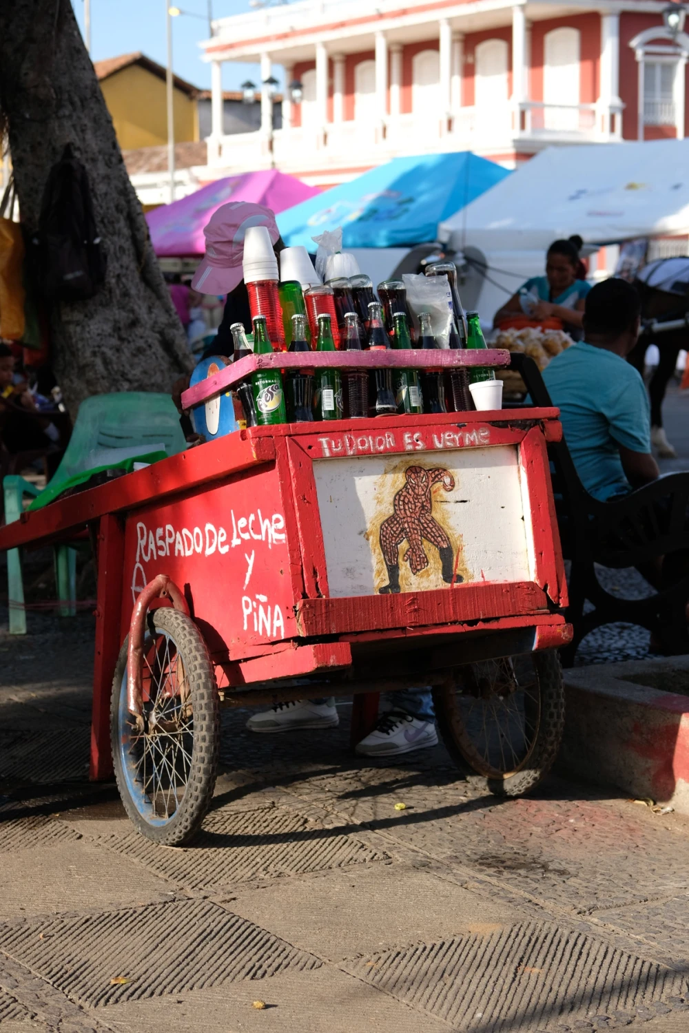 A red wooden street cart painted with a folk-art jaguar and lined with bottled drinks in a busy market area