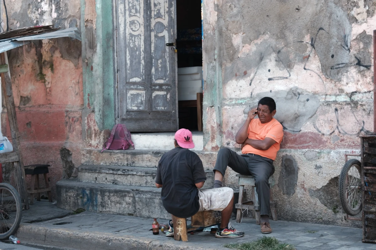 Two men sitting on the worn steps of a colonial building with peeling, distressed plaster, one playing a game on a stool