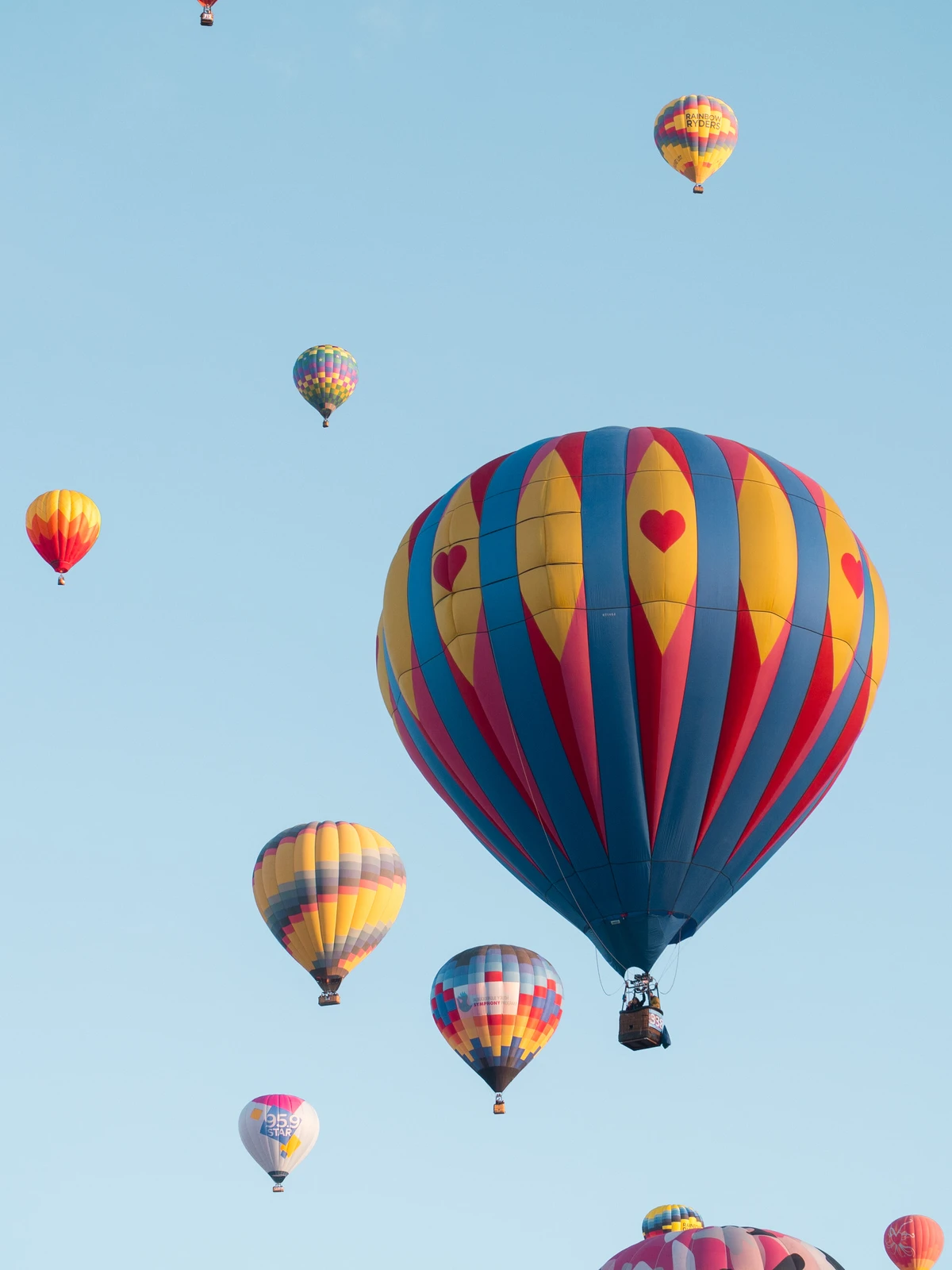 Dozens of hot-air balloons climbing into a pale blue morning sky, a blue-and-yellow balloon with red hearts among them