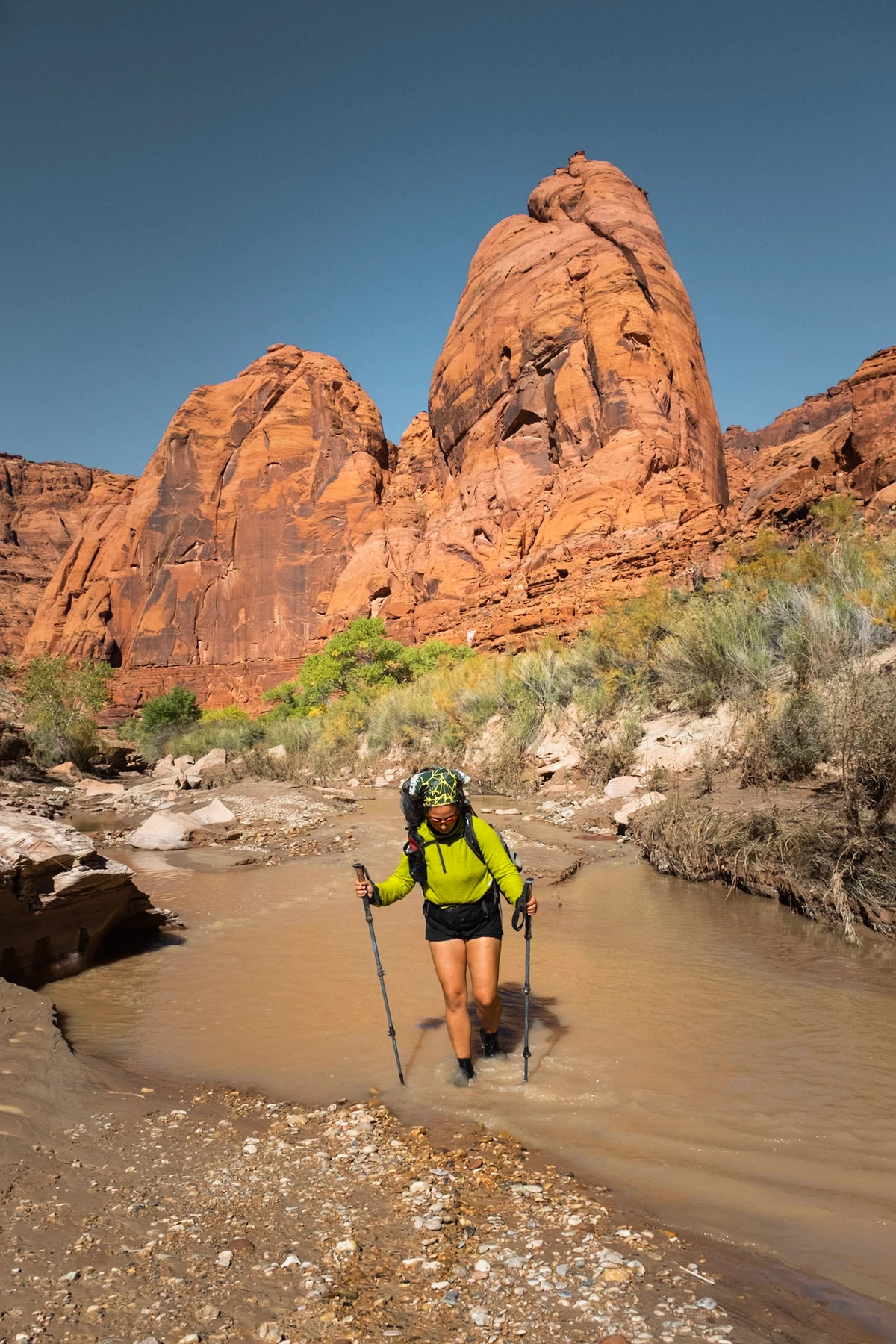 A backpacker wading a river crossing below tall red sandstone domes